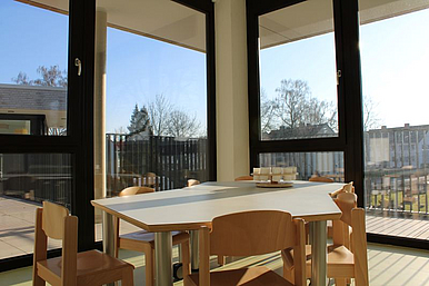 Auf dem Bild ist ein Tisch mit Stühlen zu sehen. Auf dem Tisch stehen Stifte und er befindet sich vor einer großen Fensterfront, die auf den Außenbereich blicken lässt. 