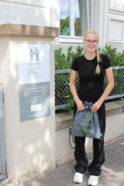 Vor dem Verwaltungsgebäude steht eine junge Frau mit einem Turnbeutel mit der Aufschrift "Ich bin einzigartig" in den Händen.