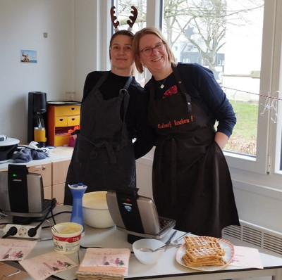 Zwei Mitarbeiterinnen stehen dicht nebeneinander und lächeln. Die linke hat ein Rentiergeweih auf dem Kopf. Vor ihenne steht ein Tisch mit Waffelteig und Waffeleisen.