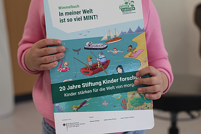 Wir sehen ein Wimmelbuch von der Stiftung Kinder forschen. Das Buch wird von einem Kind in den Händen gehalten.