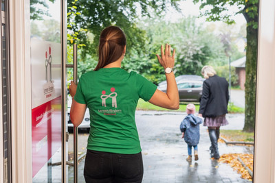 winkende Frau im grünen Shirt steht mit dem Rücken in einer Tür, 