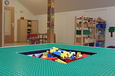 Ein Tisch auf dem Legoplatten befestigt wurden, in der Mitte des Tisches ist ein Loch in dem Legosteine aufbewahrt werden können. Im Hintergrund sind zwei Regale mit Spielmaterialien.