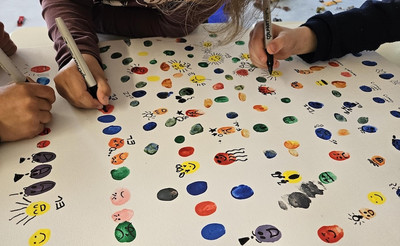 Eine Leinwand mit kleinen bunten Figuren, die unterschiedliche Gefühle zeigen. Am Rand Kinderhände, die an dem Bild arbeiten.