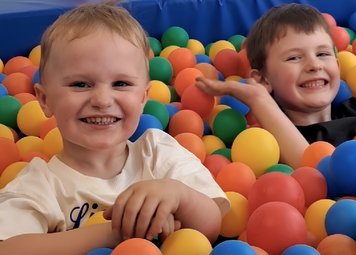 In einem bunten Bällebad sitzen zwei Kinder. Sie grinsen in die Kamera.