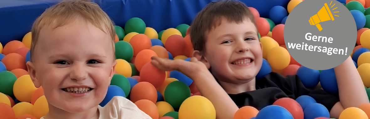 In einem bunten Bällebad sitzen zwei Kinder. Sie grinsen in die Kamera.