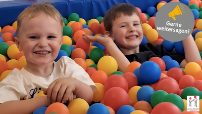 In einem bunten Bällebad sitzen zwei Kinder. Sie grinsen in die Kamera.