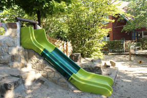 Der Spielplatz enthält viele Bäume und schattige Plätze. Ein Highlight ist die grüne Rutsche, welche im Vordergrund des Bildes ist.