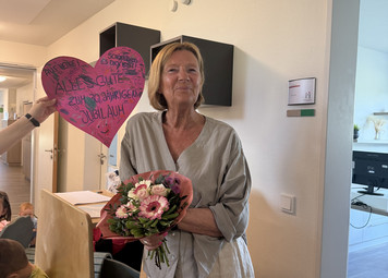Eine Frau steht lachend mit einem Blumenstrauß in einem Flur. Im Hintergrund hängt ein pinkes Herz.