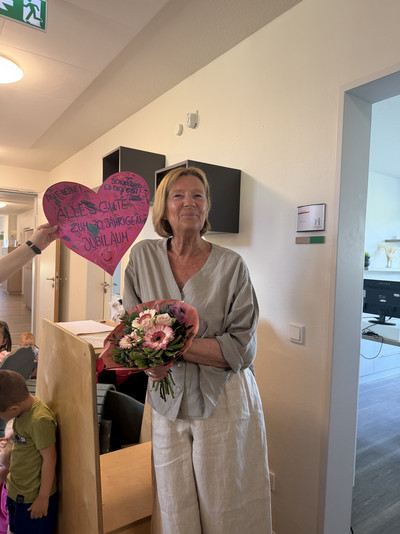 Eine Frau steht lachend mit einem Blumenstrauß in einem Flur. Im Hintergrund hängt ein pinkes Herz.
