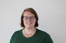 Eine lächelnde junge Frau mit kurzen dunklen Haaren. Sie trägt eine Brille und ein dunkelgrünes T-Shirt. Sie steht vor einer weißen Wand.