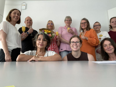 Drei Frauen hocken hinter einem Tisch, sieben weitere Personen stehen mit erhobenen Gläsern und einem Blumenstrauß um die drei herum