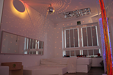 Eine bunte Säule sieht man vorne rechts, eine Discokugel hängt an der Wand, Ein Spiegel hängt links an der Wand. Eine gemütliche Ecke ist links im Hintergrund zu erkennen