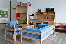 Eine Puppenecke mit einer Küche an der Wand, daneben steht ein großes regal mit Materialien ind einem Kinderbett. Davor steht die Sitzmöglichkeit 