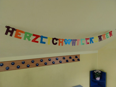 bunte Girlande mit Herzlich willkommen vor gelber Wand, darunter leere Garderobenhaken