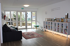 Ein Bücherreagl rechts an der Wand, links ein großes Sofa und in der Mitte sieht man einen Teppich, sowie eine Tür die zum Außengelände führt