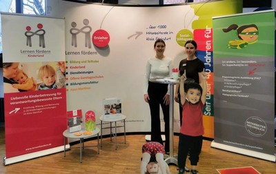 Messestand mit Aufstellern und Flyern. In der Mitte zwei Mitarbeiterinnen und zwei Kinder-Aufsteller