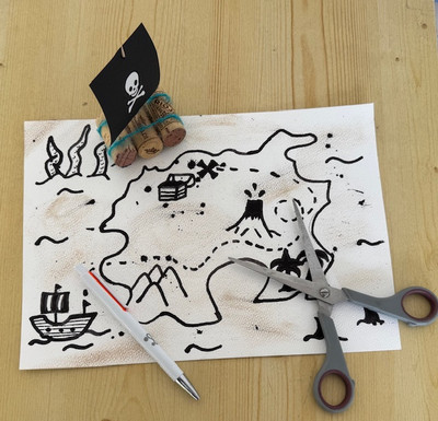 Eine Piratenschatzkarte auf einer hölzernen Untergrund. Die Karte zeigt eine Langfläche, ein Schiff und Wellen. Unten am Rand liegt eine Schere, sowie ein Kugelschreiber.