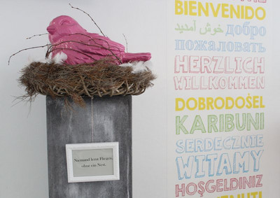 Hier sieht man einen pinken Vogel in einem Nest auf einem Podest. Ein Bilderrahmen hängt vor dem Podest mit dem Spruch: "Niemand lernt fliegen ohne ein Nest."