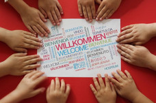 Ein Schild auf dem in vielen verschiedenen Sprachen "Willkommen" steht. Das Schild wird von vielen Kinderhänden eingerahmt.