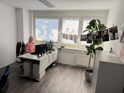 Eine Frau sitzt an einem Schreibtisch mit Computer in einem Büro, in dem die Wäscheleine mit den Fotos hängt
