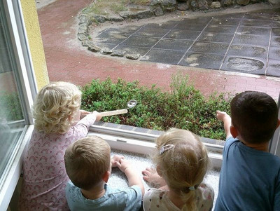 Vier Kinder stehen vor einem geöffnetem Fenster. Die Steine und Pflanzen draussen vor dem Fenster sind nass vom Regen. Ein Kind hält eine Suppenkelle mit gestrecktem Arm und versucht in dieser Regen zu fangen.