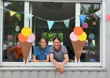 Zwei Frauen schauen lächelnd aus einem Fenster. Links und rechts am Rahmen hängt ein Eis im Hörnchen aus Pappe. Bunte Wimpel hängen geschwungen über der Fensteröffnung.
