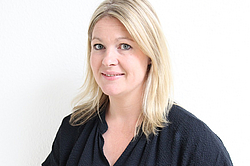 Eine lächelnde Frau mit blonden Haaren vor einer weißen Wand. Sie trägt eine dunkelblaue Bluse.