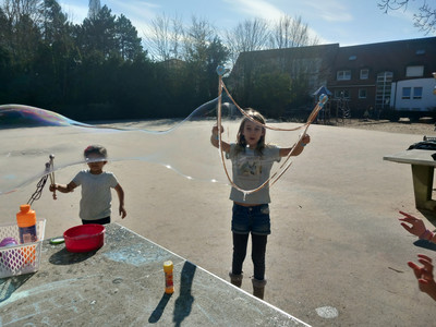 Zwei Kinder auf dem Pausenhof der Schule. Sie spielen mit Seifenblasen. Vor ihnen auf einer Tischtennisplatte kleine Seifenblasenpackungen.