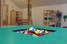 Ein Tisch auf dem Legoplatten befestigt wurden, in der Mitte des Tisches ist ein Loch in dem Legosteine aufbewahrt werden können. Im Hintergrund sind zwei Regale mit Spielmaterialien.