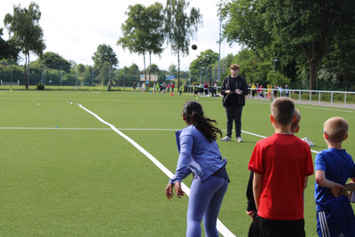 Vier Kinder stehen auf einem Rasenplatz, ein Mädchen wirft einen Ball. Im Hintergrund steht ein Junge und schreibt die Ergebnisse vom Wurf auf. 