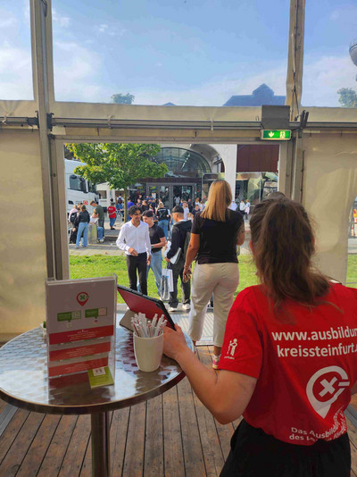 Im Hintergrund der Eingang zum Messezelt, im Vordergrund eine Frau in rotem T-Shirt vom Local X-perts Messe-Team.