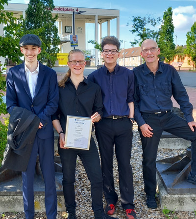 Auf dem Foto sind vier lächelnde Personen zu sehen. Die Person sind von links betrachtet David Piesik, Nils Zimny, Pascal Esser und Michael Rastbichler. Die zweite Person von links hält eine Auszeichnung als Prüfungsbester in der Hand. 