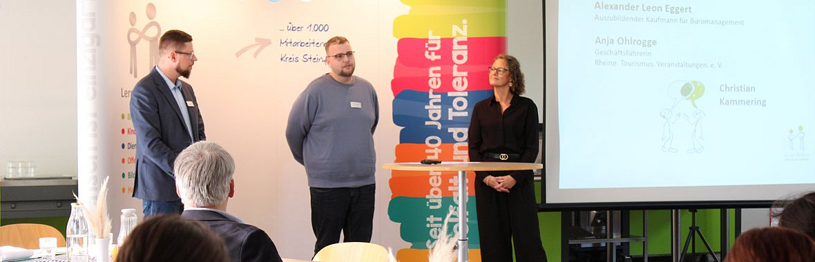 vorne Zuschauer im Hintergrund drei Menschen im Gespräch vor einer Werbewand 