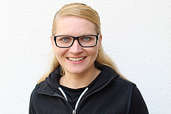 Eine lächelnde junge Frau mit halb zurückgebundenen blonden Haaren und Brille. Sie steht in einer dunklen Jacke vor einer weißen Wand.
