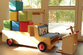 Ein heller Raum mit einem Holz-Auto ist zu sehen. Beladen ist das Auto mit verschiedenen Stoffbausteinen und lädt die kleinsten Kinder zum "Fahren" ein.
