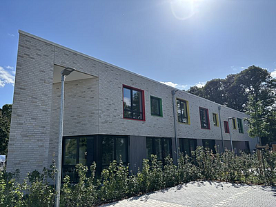 Auf dem Bild sieht man die Kinderland Einrichtung, ein beiges Gebäude mit bunten Fensterlaibungen. 