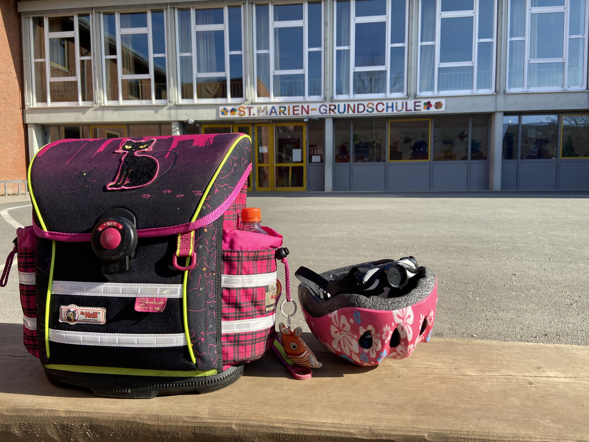 In der linken Ecke eine pink schwarze Schultasche mit einem Fahrradhelm auf einer Steinmauer. Im Hintergrund der Eingang der Marienschule mit einer großen Fensterfront.