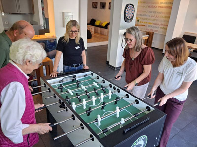 vier Personen spielen an einem Kicker, eine Person sieht zu