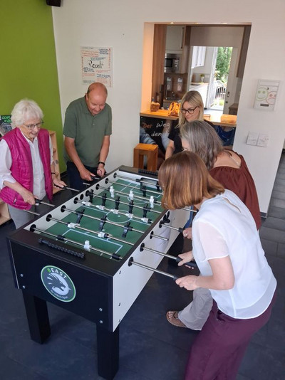 vier Personen spielen an einem Kicker, eine Person sieht zu