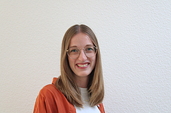 Eine lächelnde junge Frau mit Brille, mittellangen blonden Haaren und einer orangen Jacke. Sie steht vor einer weißen Wand.