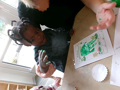 Ein Kind mit grüner Fingerfarbe an der Hand für einen Handabdruck auf Papier der auf dem Tisch vor dem Kind liegt