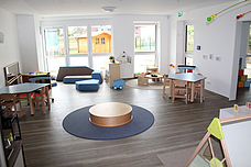Ein heller Gruppenraum. Mit Tischen und Stühlen, einem Sofa, einer Puppenecke rechts in der Ecke und einem Podest in der Mitte, auf einem blauem Teppich 