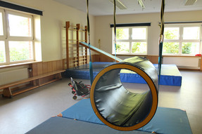 Eine große Turnhalle mit Bewegungsmöglichkeiten. In der Mitte der Turnhalle schwebt eine große Rolle, die Kinder zur Bewegung animieren soll.