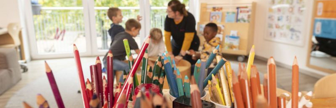 Im Vordergrund Buntstifte, dahinter vor einer Fensterfront eine Gruppe spielende Kinder mit einer Erzieherin in der Mitte.
