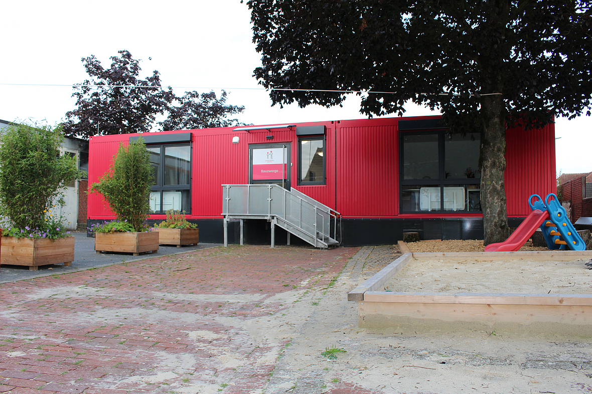Eine Außenansicht der Einrichtung. Ein roter Containerbau, umgeben von Bäumen und Grünpflanzen. Davor ein Spielplatz mit einer Rutsche.