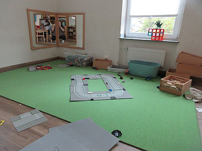 Man sieht auf dem Bild die Ecke eines Raumes. Auf dem Boden liegt ein hellgrüner Teppich. Auf dem Teppich sind verschiedenen Spielzeuge verteilt. An der Wand hängt ein Spiegel, zudem erhellt ein Fenster den Raum.