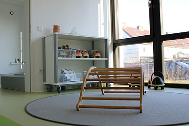 Auf dem Bild ist ein Kletterbogen zu sehen im Vordergrund, welcher auf einem runden Teppich vor einer Fensterfront steht. Hinter dem Bogen ist ein großes offenes Regal mit Bewegungsmaterialien. 