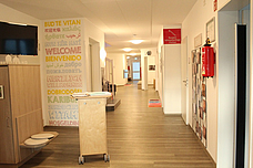 Der helle Eingangsbereich. An der linken Seite hängt der Bildschirm, daneben steht das Begrüßungspult mit der Wand wo "Herzlich Willkommen" auf verschiedenen Sprachen steht. Rechts ist die Team-Wand zu sehen und den Eingang des Büros. 