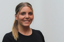Eine lächelnde junge Frau mit dunkelblonden Haaren, die zu einem Zopf gebunden sind. Sie steht vor einer weißen Wand und trägt ein schwarzes T-Shirt.