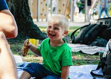 Wir sehen einnen Jungen, auf einer Picknickdecke sitzen. Er lacht und hält eine Brezel in der Hand.
